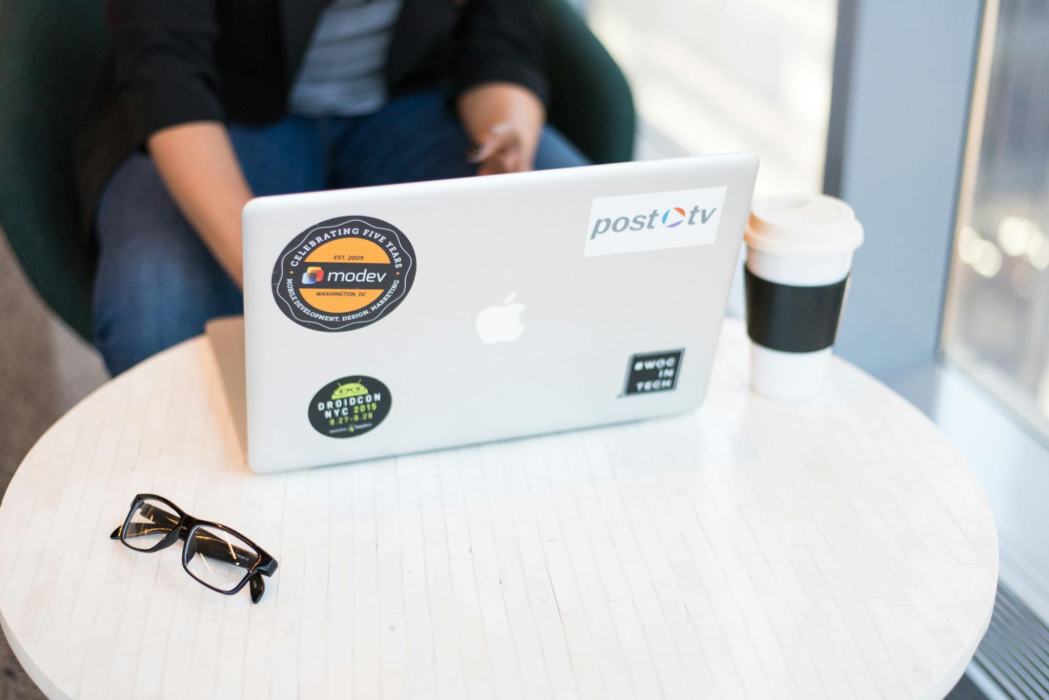 Person sitting indoors working on a laptop with stickers, coffee, and eyeglasses on a table.